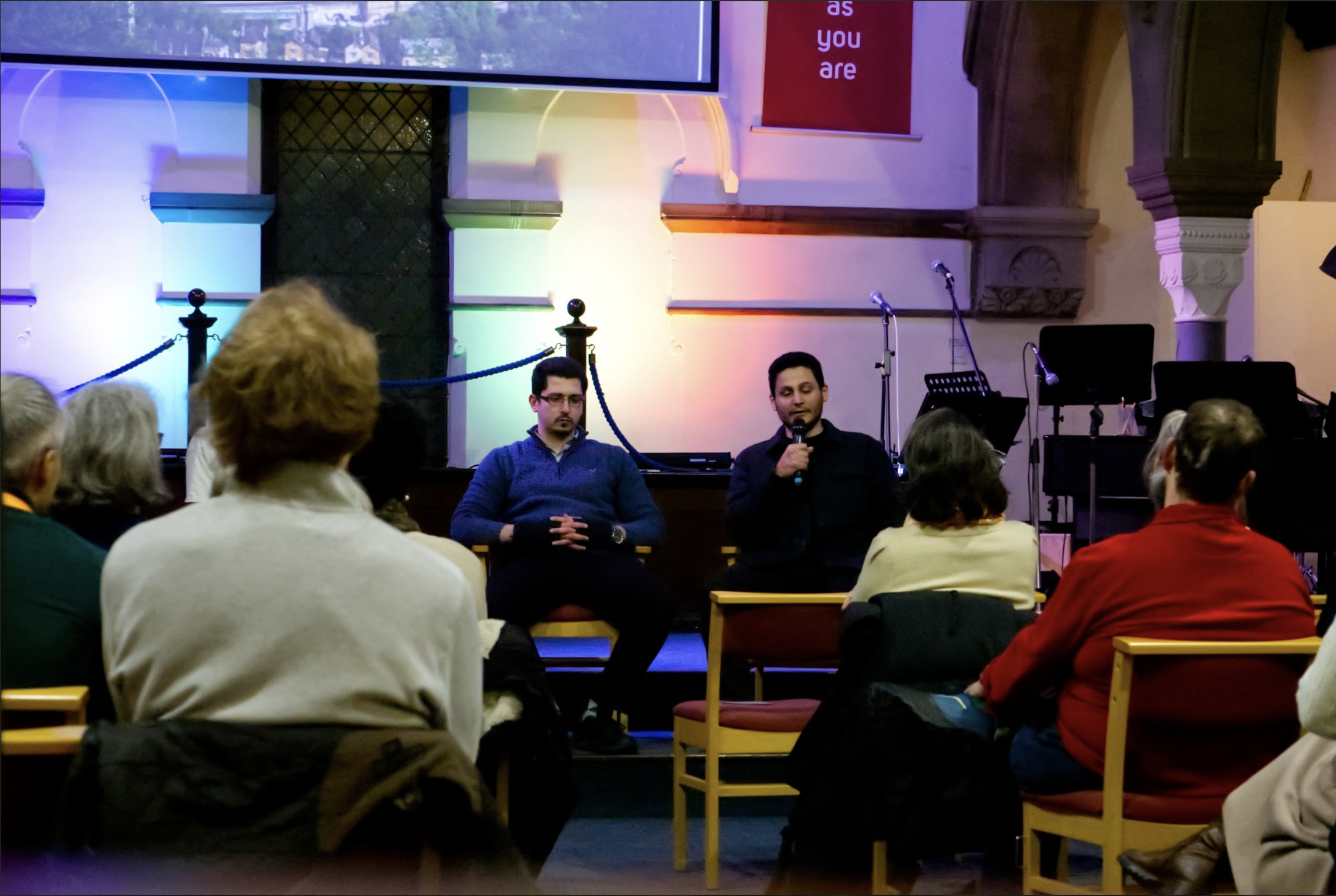 Photo of two people sitting in chairs on a stage, one is talking into a microphone
